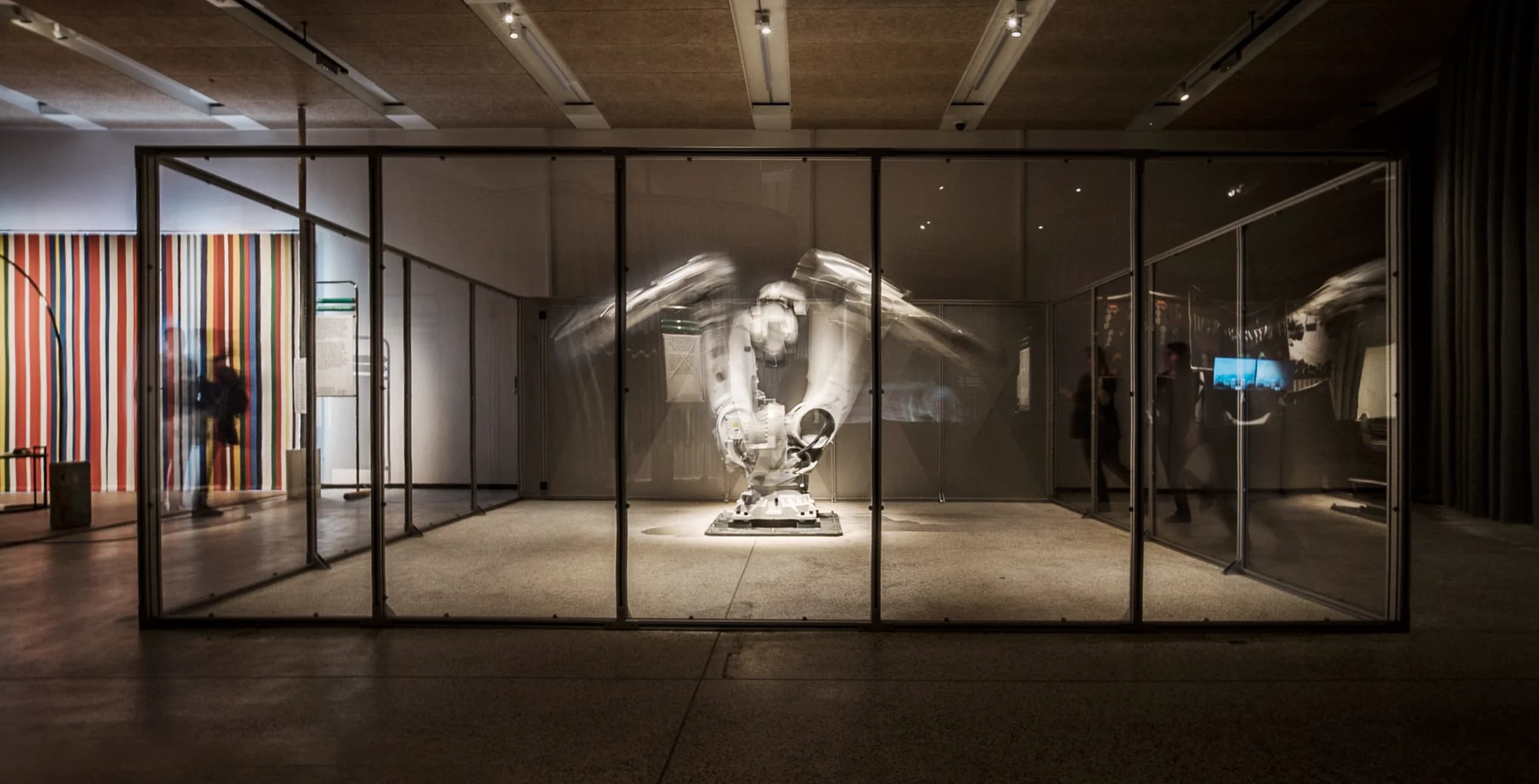 A blurred, moving robotic arm is displayed in a glass enclosure. A colorful striped wall and silhouetted figures are visible in the background.