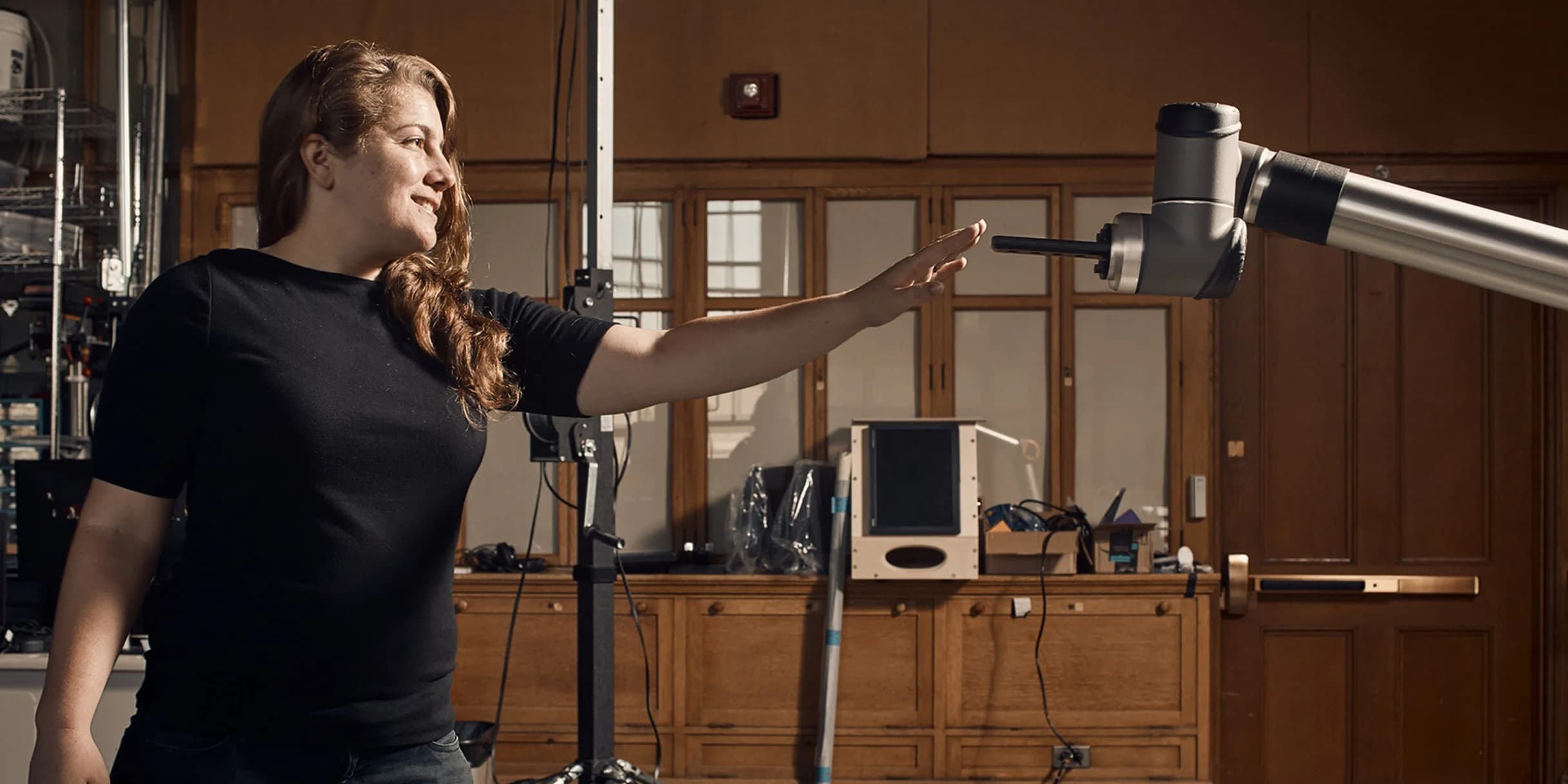 A woman reaches toward a robotic arm extending from the right side of the photo. She wears a black shirt and smiles.