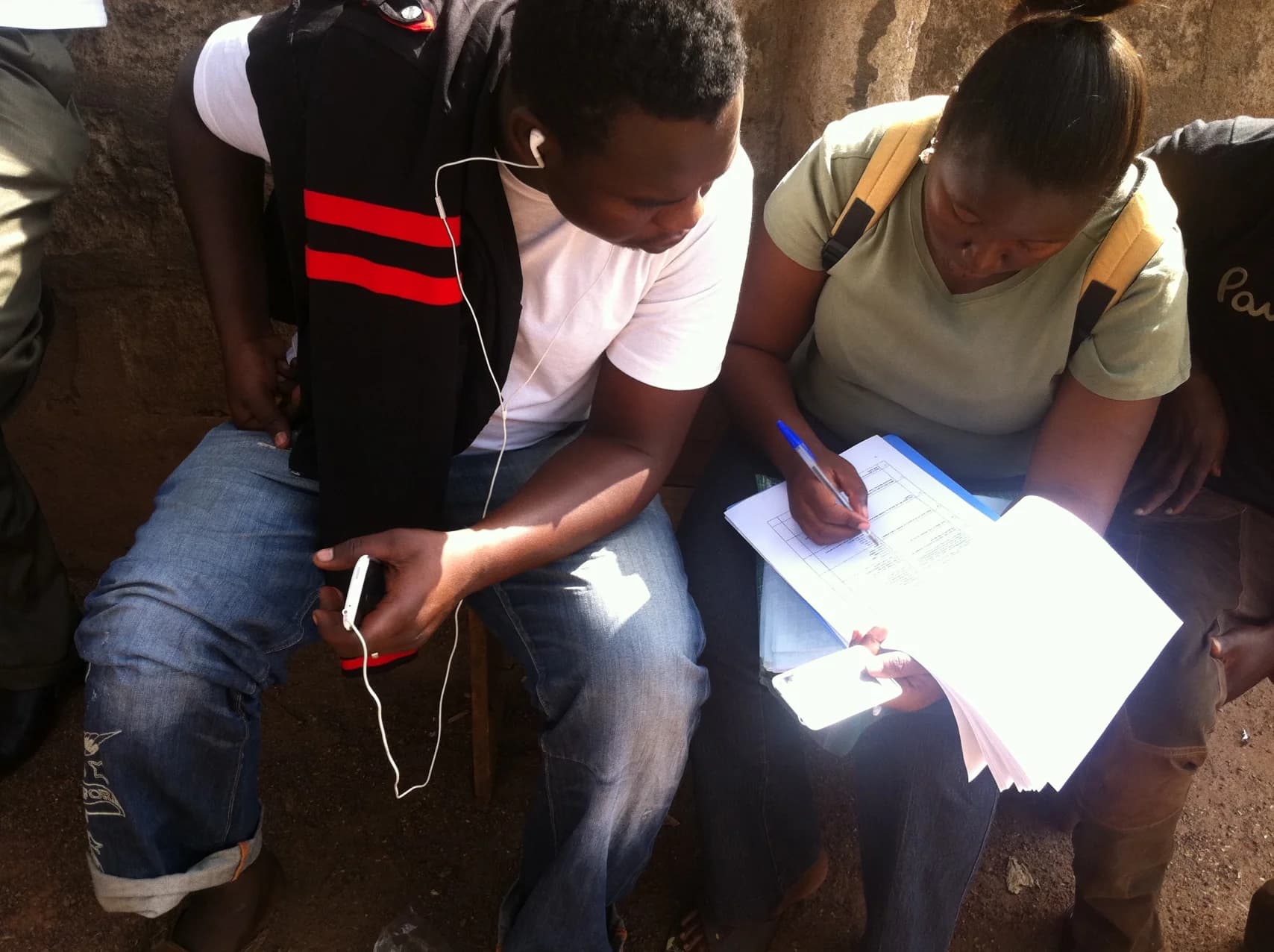 Two people sit, collaborating on a clipboard document. One writes with a pen; the other holds a phone.