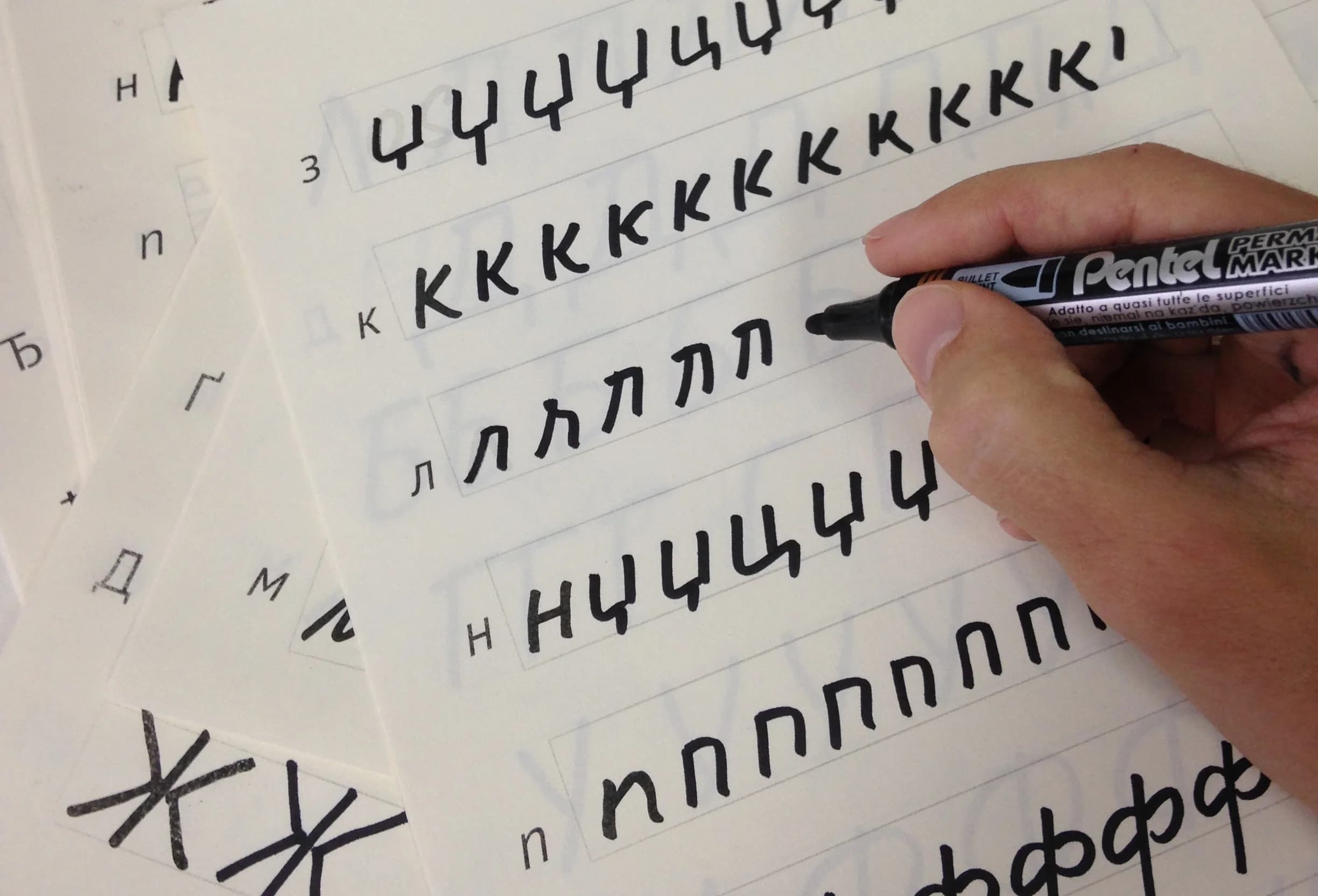 A hand writes cursive Cyrillic letters with a black marker on lined paper. Other practice sheets are visible beneath.
