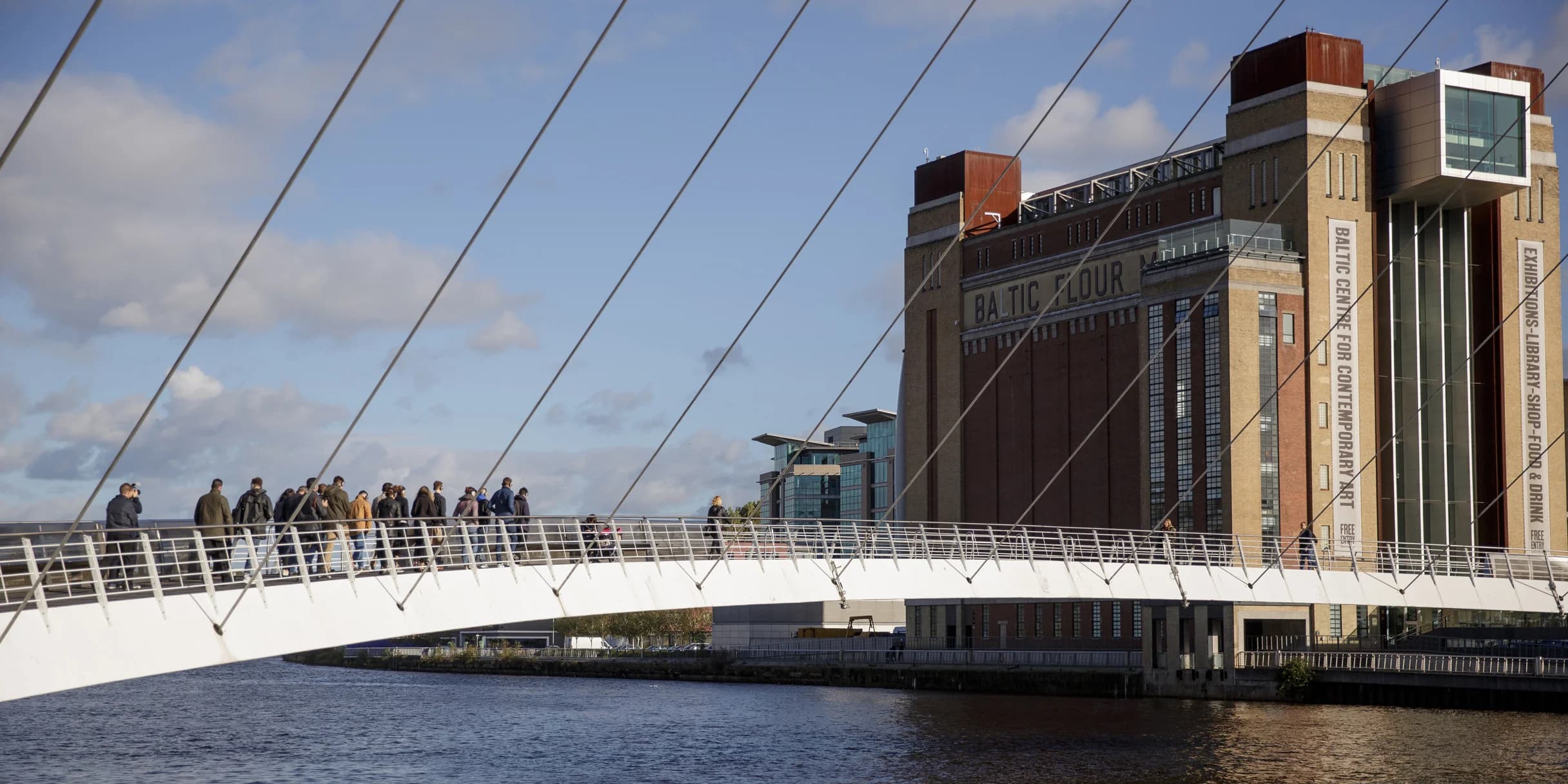 People walk across the Gateshead Millennium Bridge. The contemporary art museum, Baltic Centre for Contemporary Art, stands in the background.