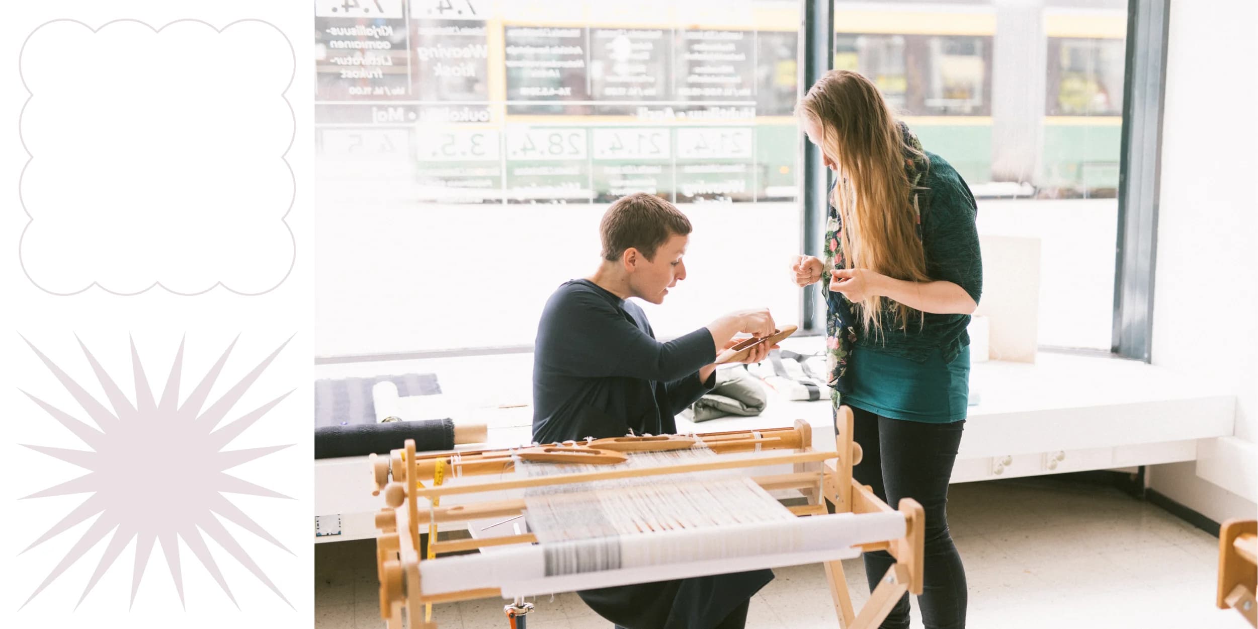 Two people work at a small loom in a brightly lit room with large windows and rolls of fabric nearby. One person sits at the loom, and the other stands beside them, assisting.