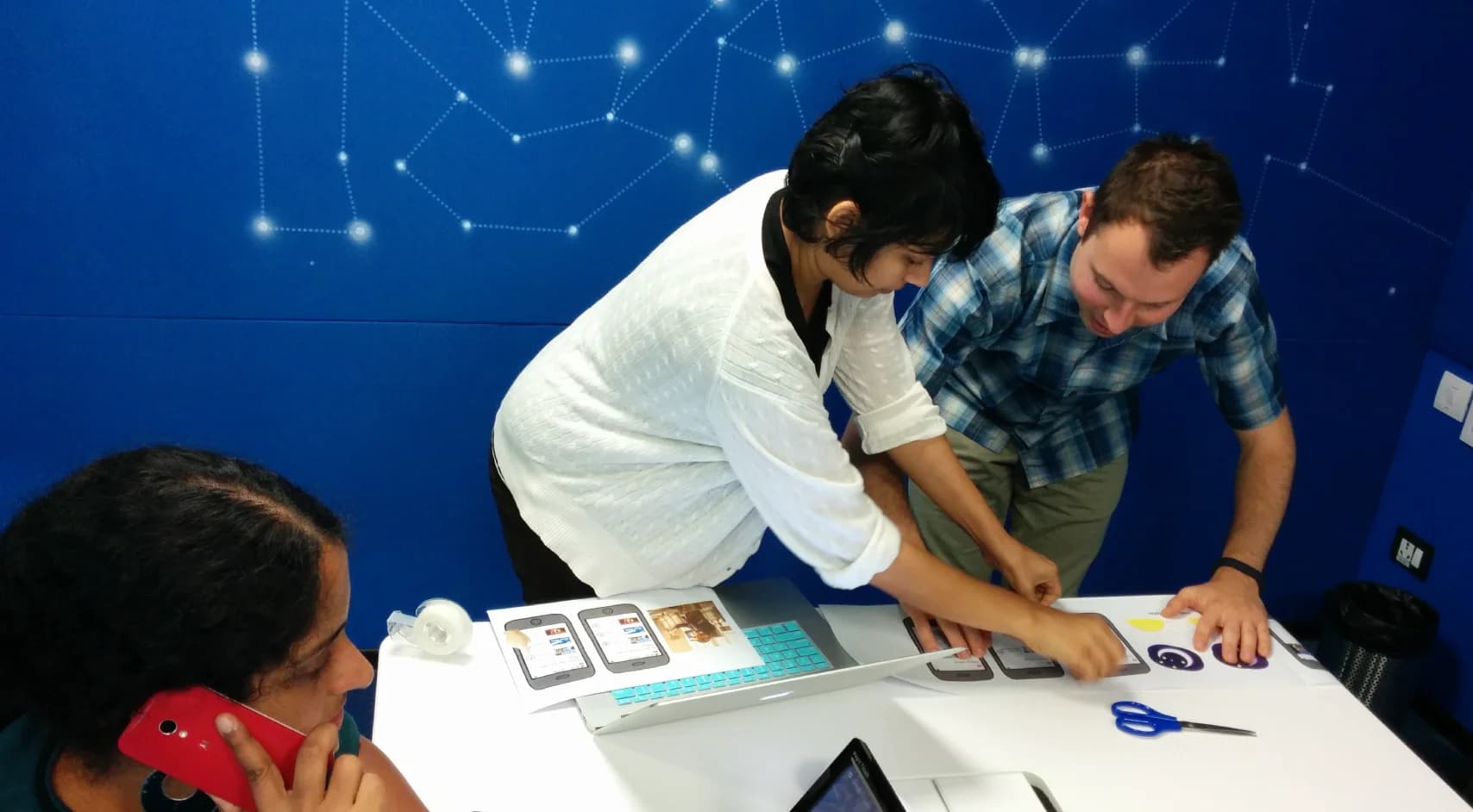 Three people gather around a white table, collaborating on printed mobile app designs. Two stand, one sits while talking on a red phone. A laptop and scissors rest on the table.