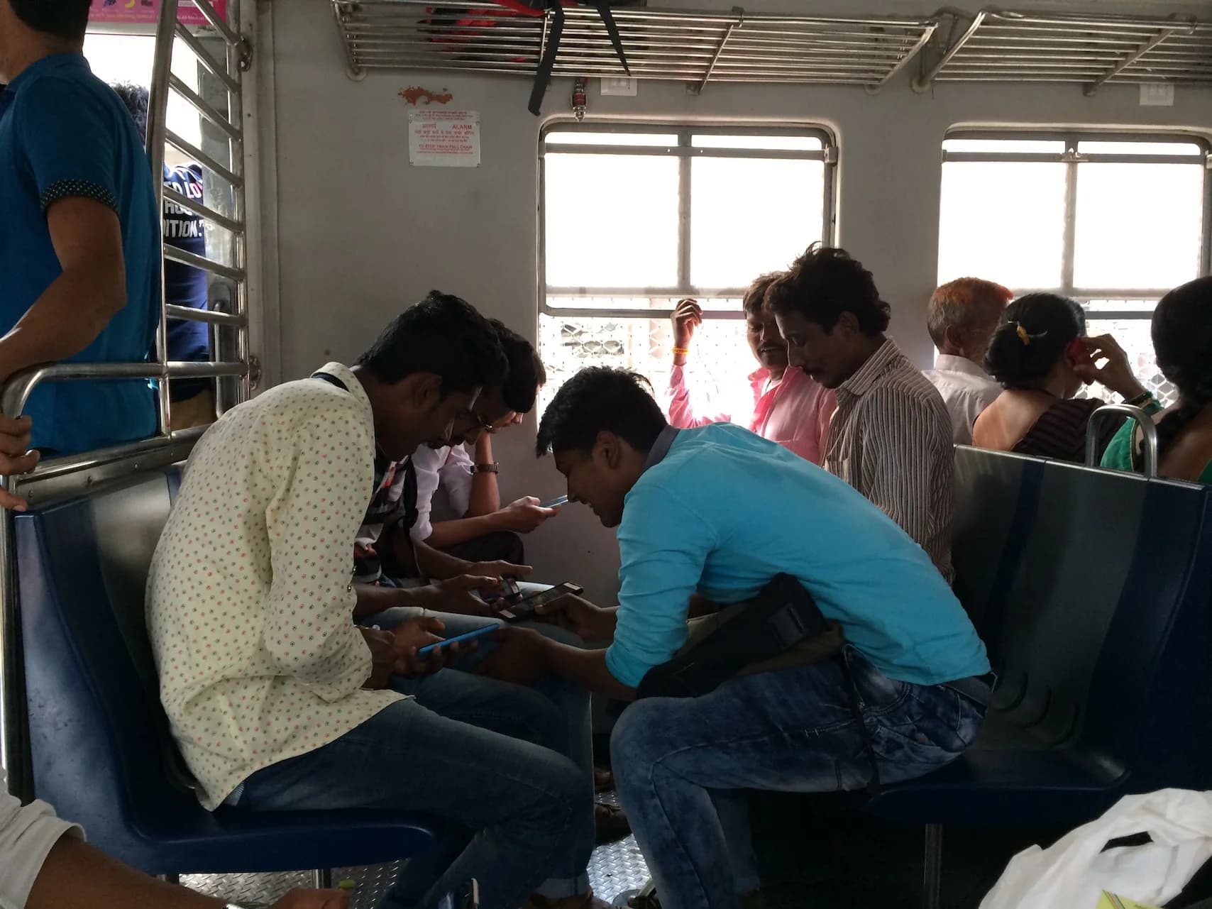 Several seated men look at smartphones on a crowded commuter train. Others passengers behind them look out the window or rest.