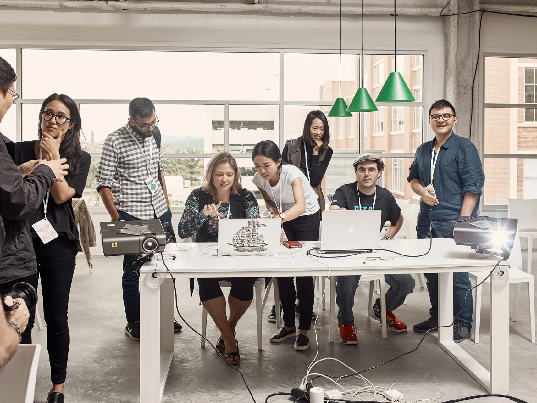A diverse group collaborates around a white table, using laptops and projectors in a bright, modern workspace.