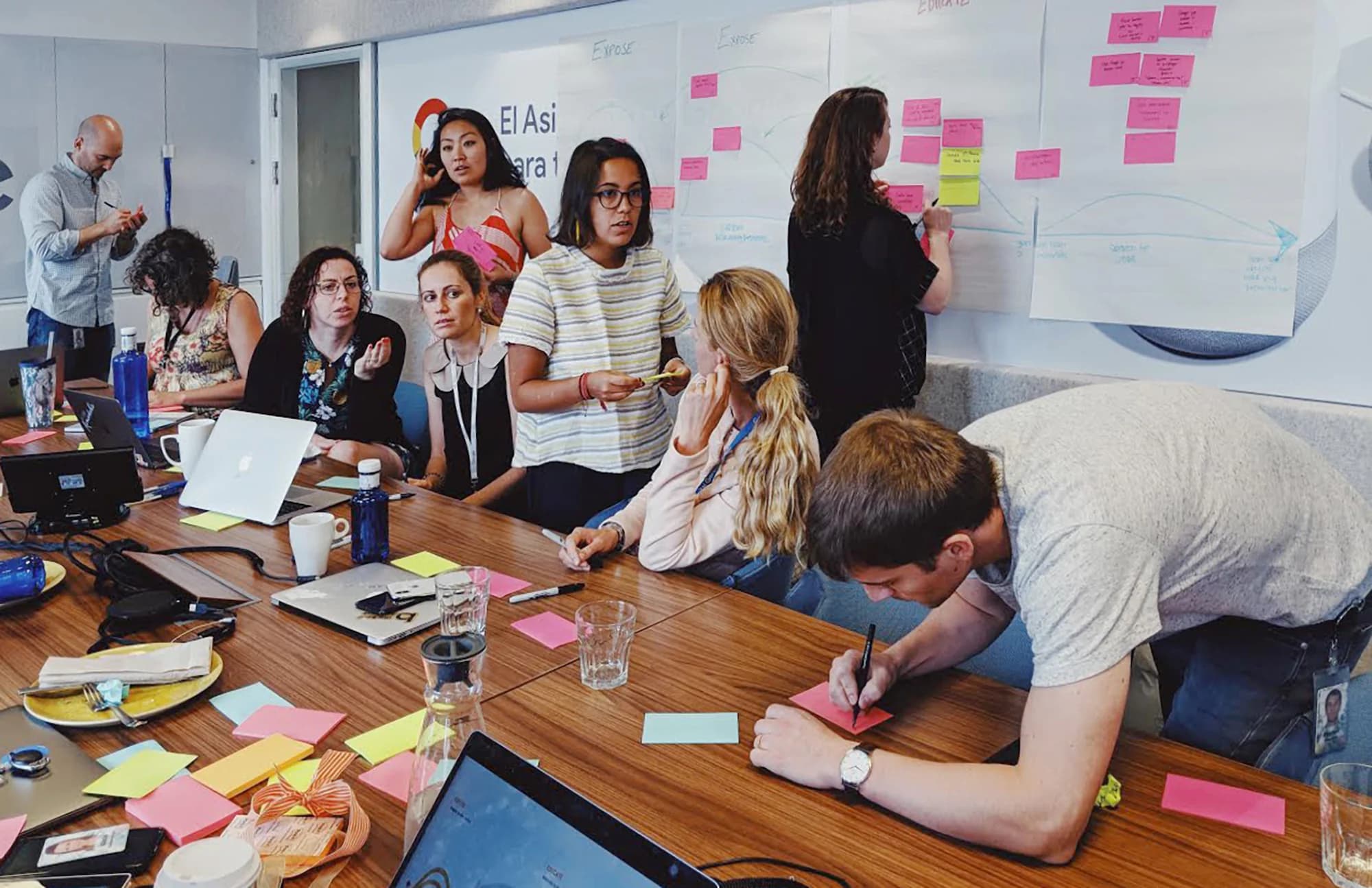 A team collaborates around a table, using laptops and sticky notes on a whiteboard during a workshop.