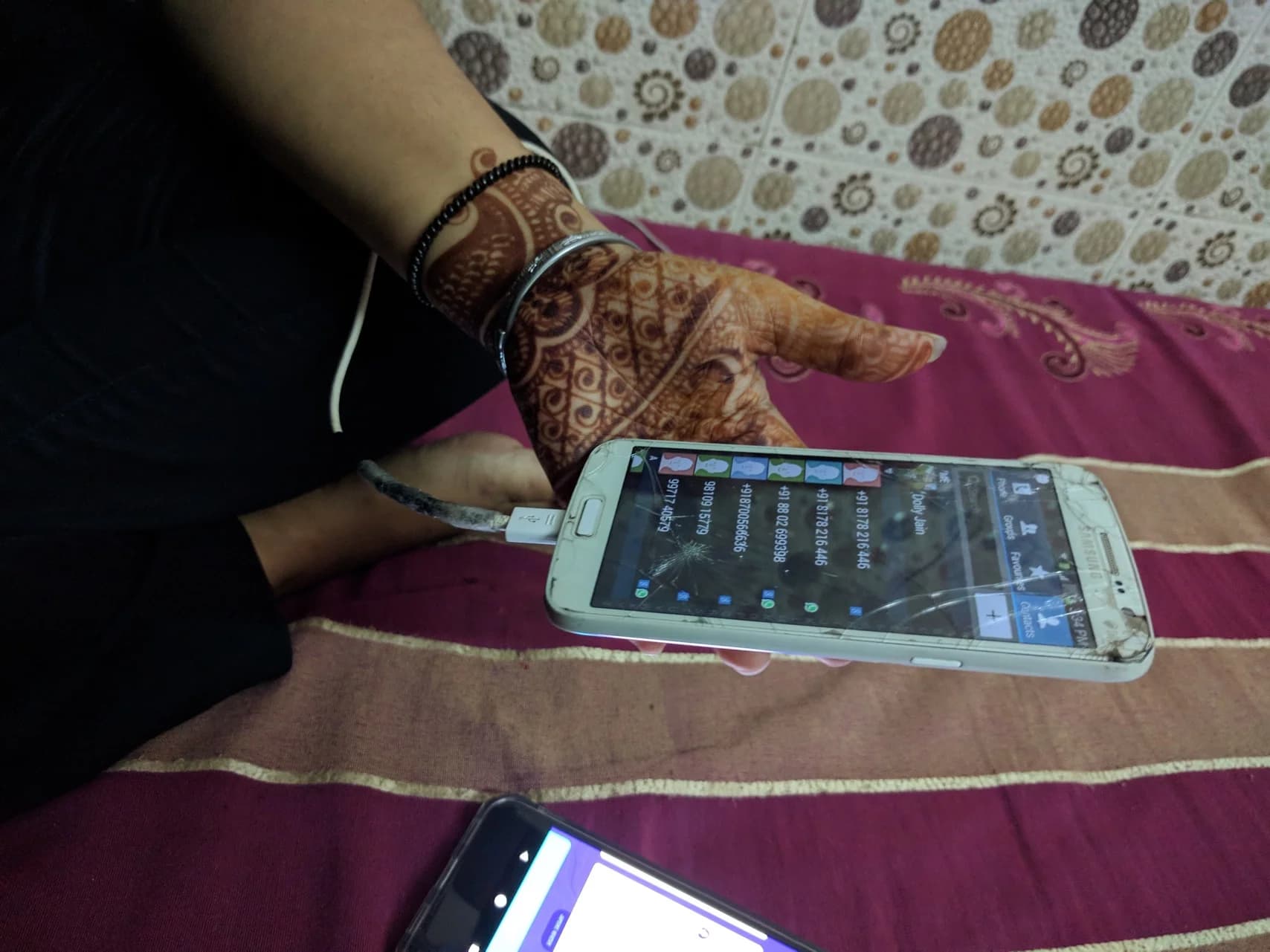 A hand with henna holds a cracked white Samsung phone plugged into a charging cable.