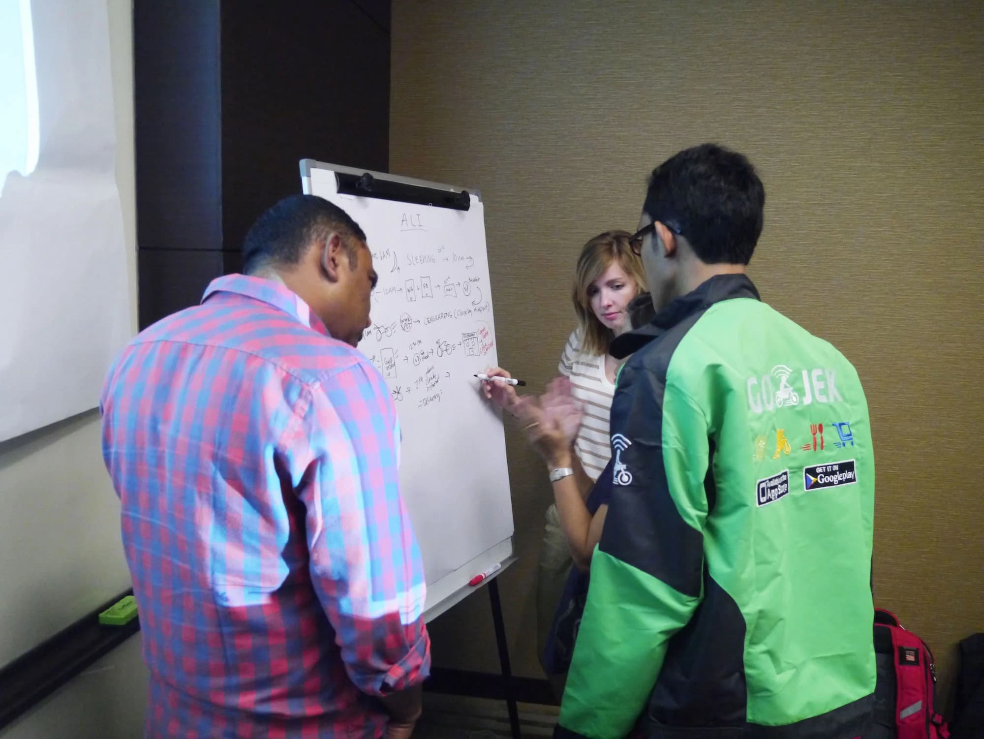 Three people collaborate on a whiteboard. One person writes a schedule, another claps, and a third wears a bright green Gojek jacket.