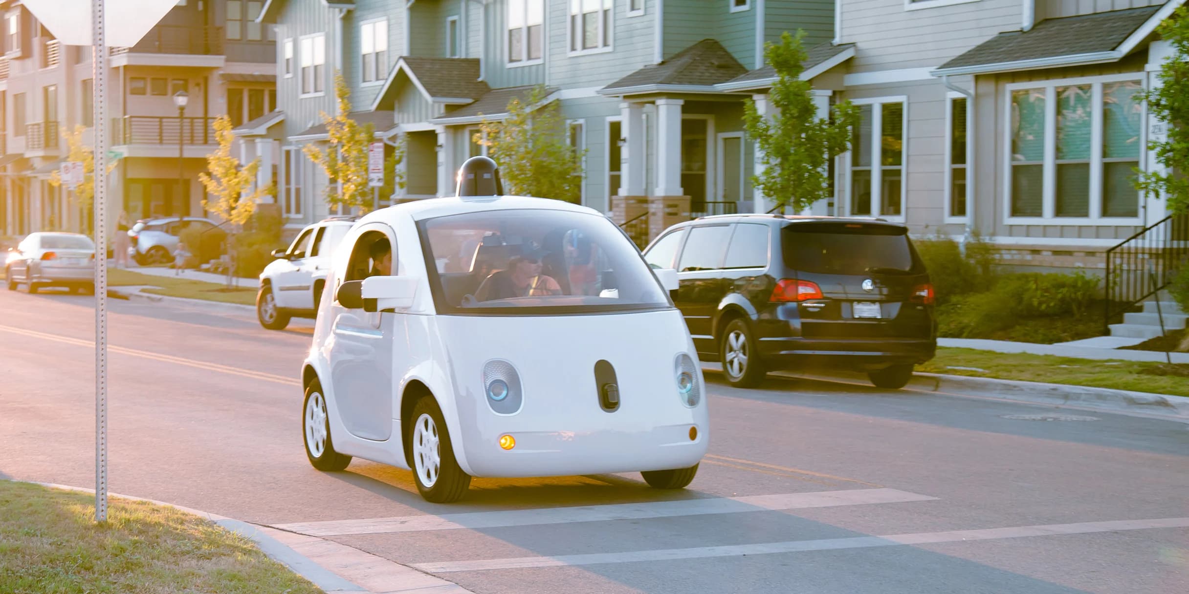 A white, two-passenger autonomous Waymo car drives down a neighborhood street at sunset.