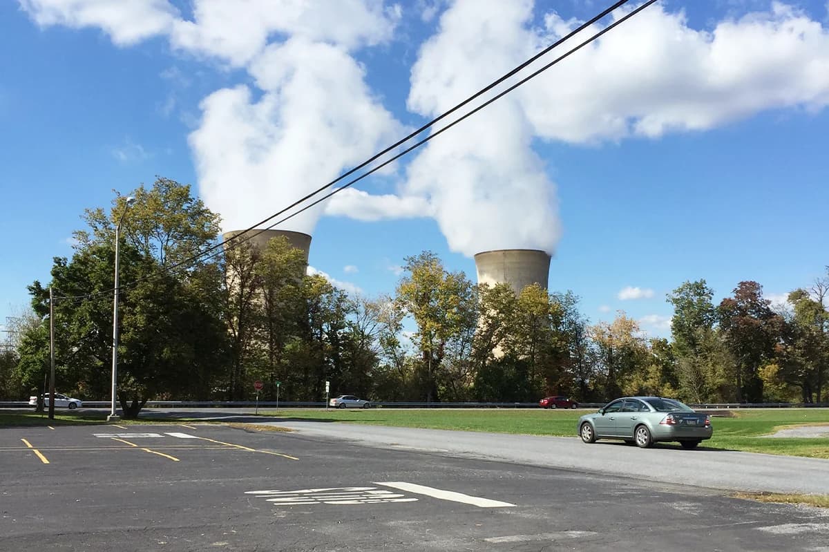 Two large cooling towers emitting steam rise above trees near a parking lot and road.
