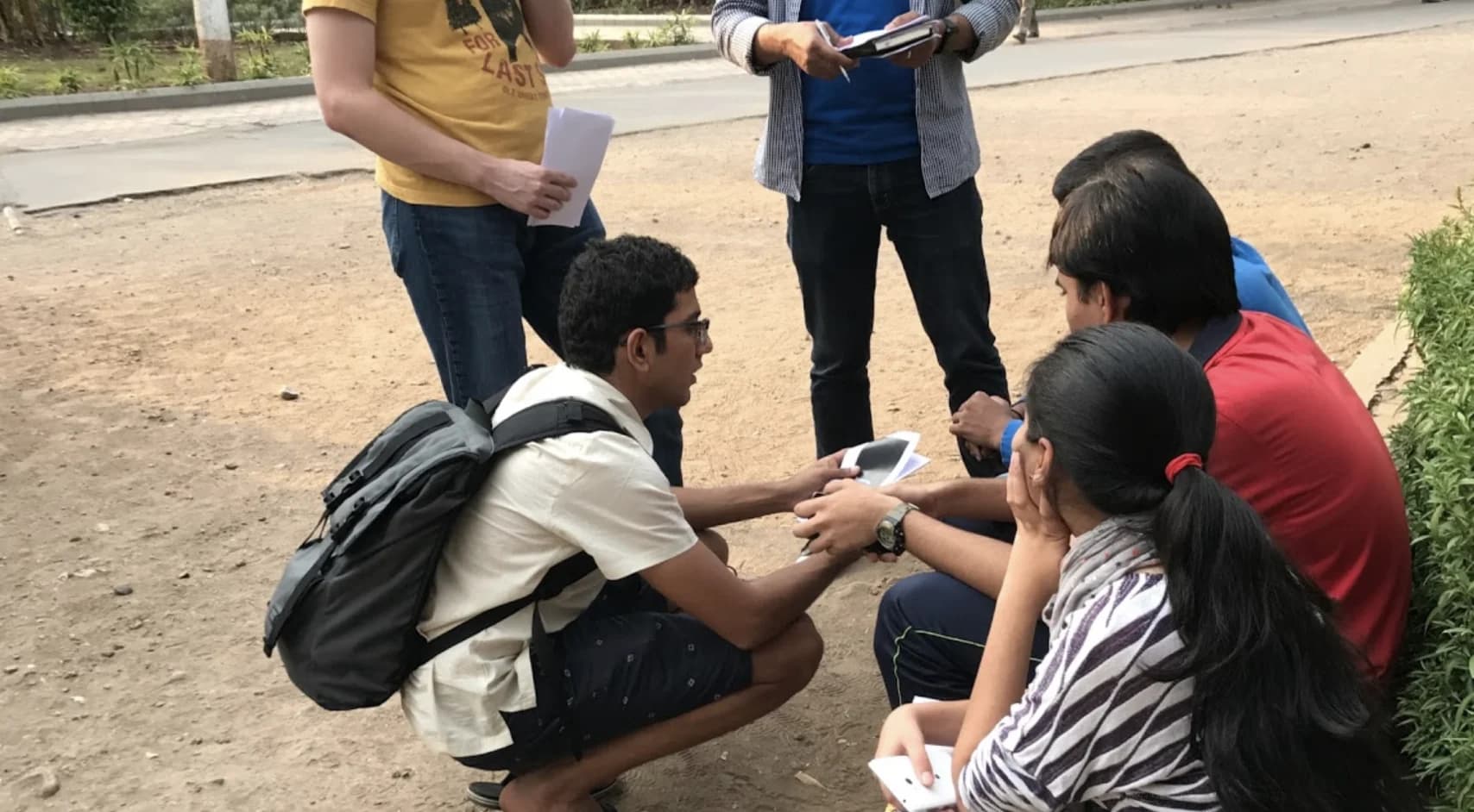 Five people collaborate outdoors. Two look at papers, while three review information on a small tablet.