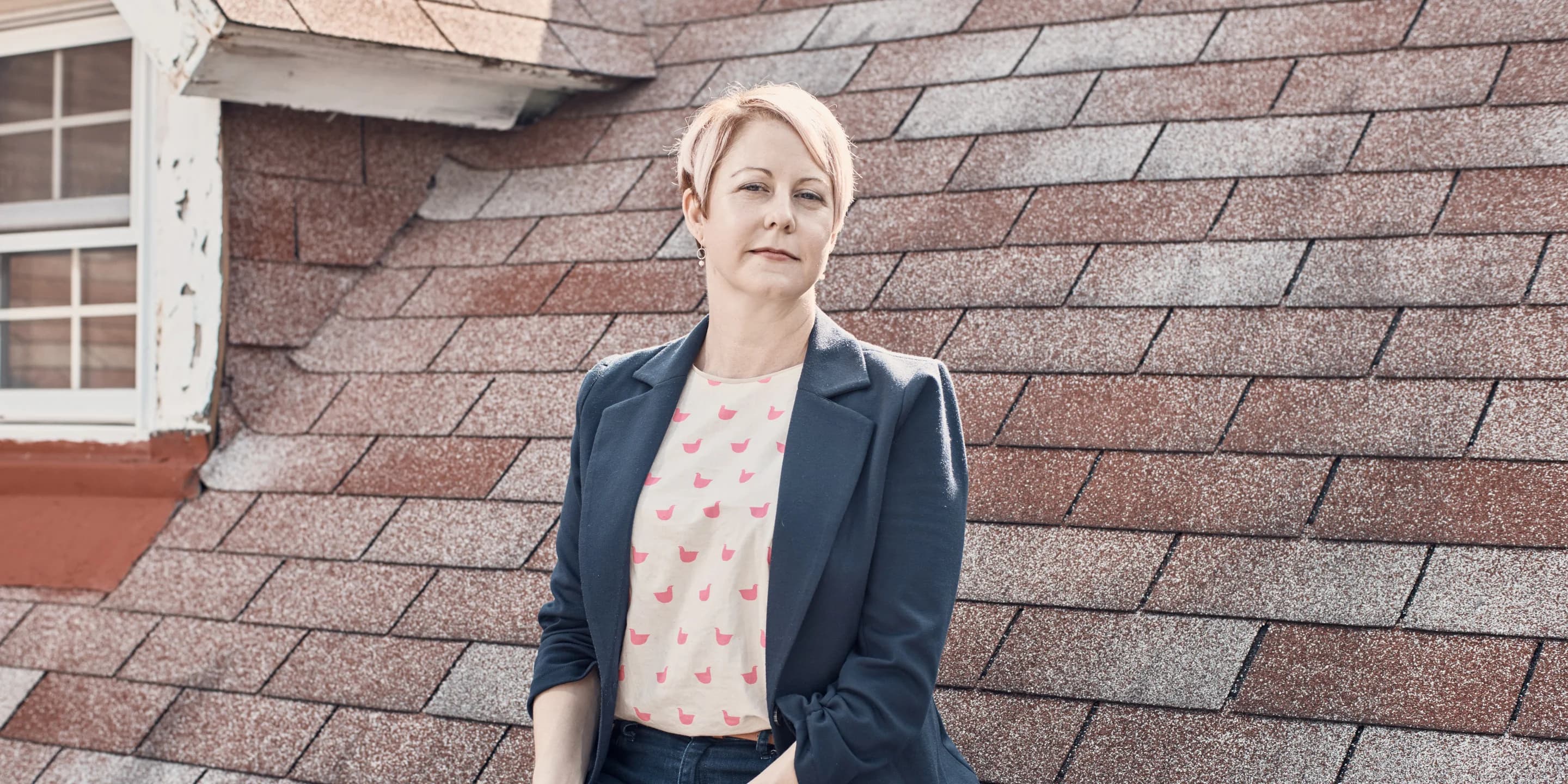 Confident person with short pink hair and a dark blazer stands before a shingled roof.