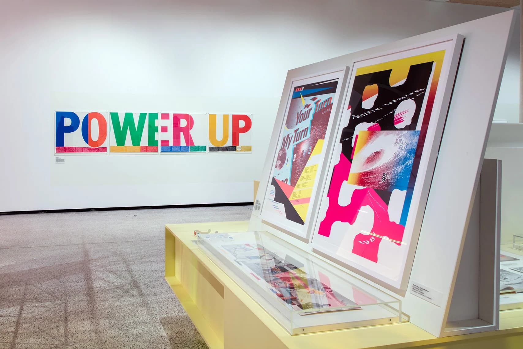 Bold, colorful graphic posters are displayed on a wall and in a display case in a gallery setting.