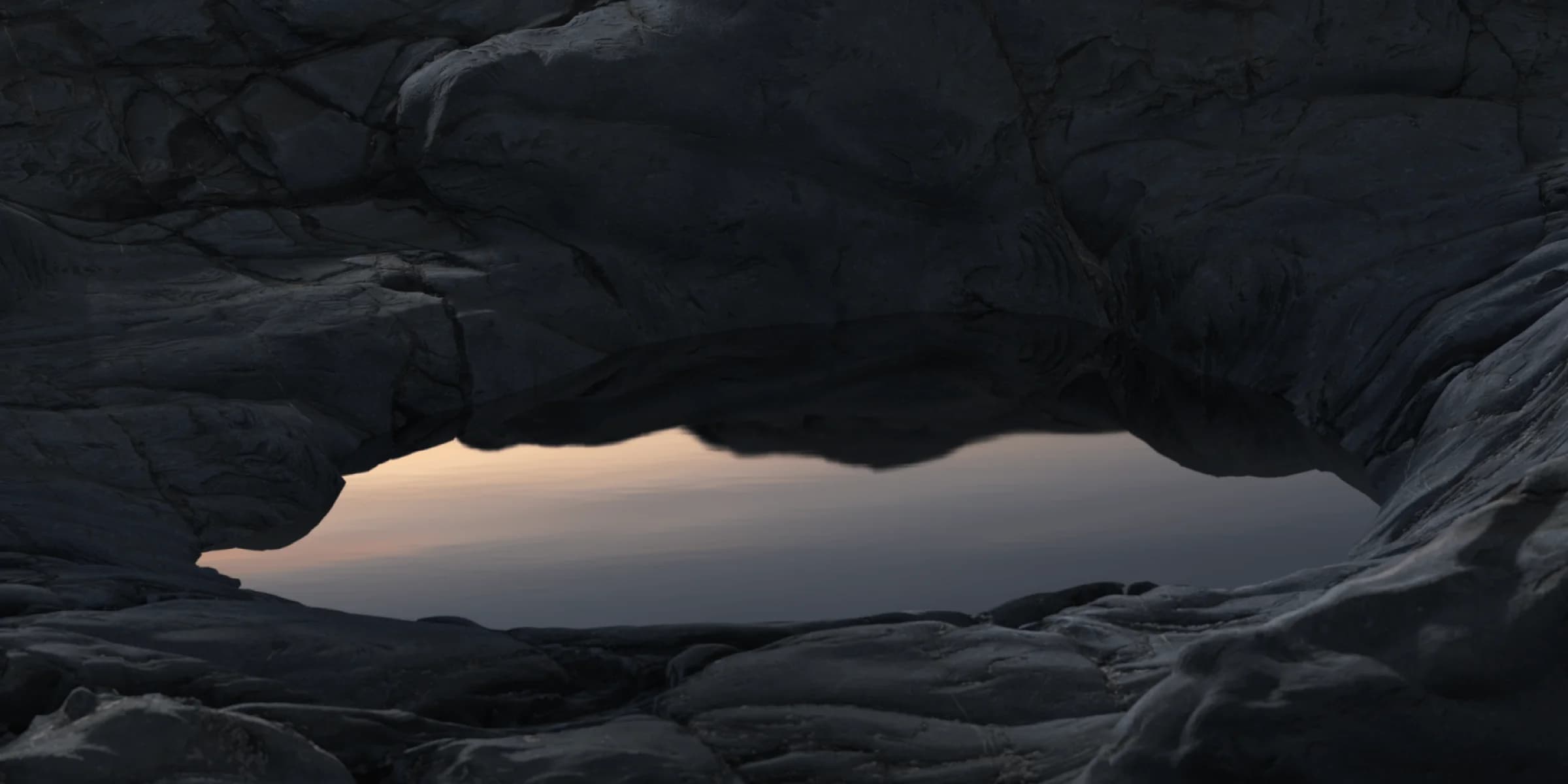 Dark, craggy rocks surround a calm, reflecting pool. A soft, pinkish sunset peeks through the opening.
