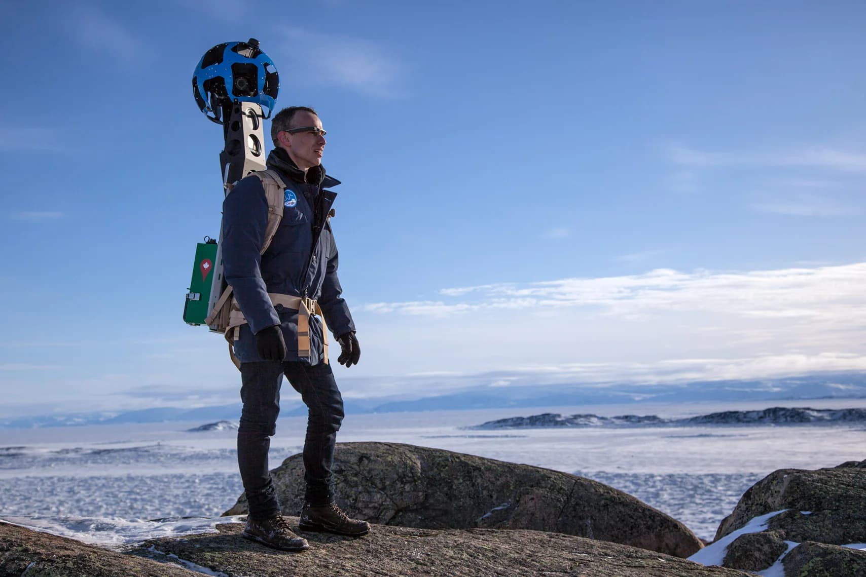 Man with a 360-degree camera backpack stands on rocks overlooking a frozen, snowy landscape.