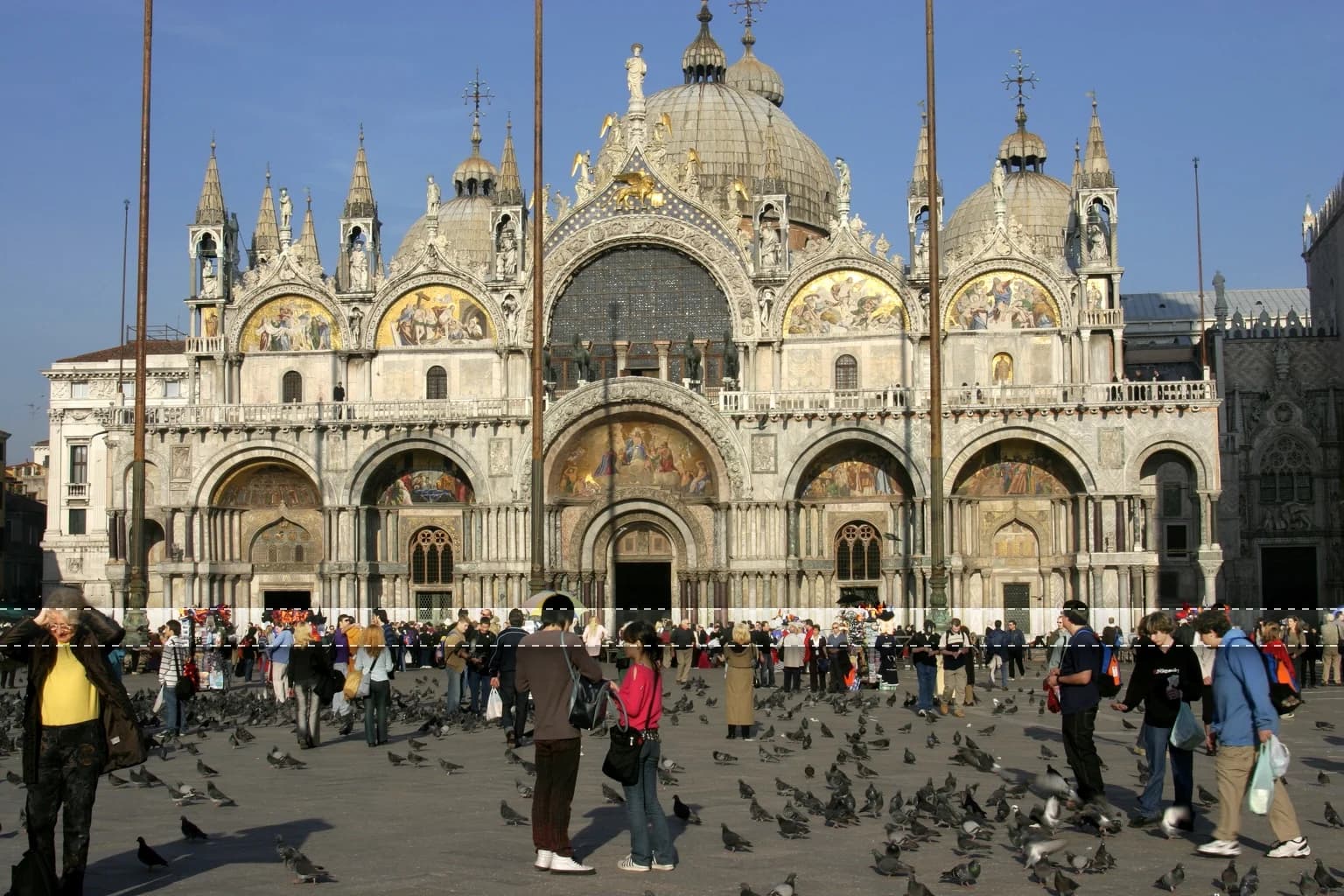 St. Mark's Basilica, Venice, with its ornate facade and domes, towers over a plaza filled with people and pigeons.