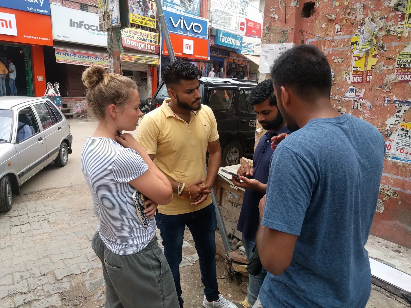 Four people gather on a bustling city sidewalk in India. Three men look at a phone, while a woman with a notebook observes. Storefronts and colorful advertisements line the street.