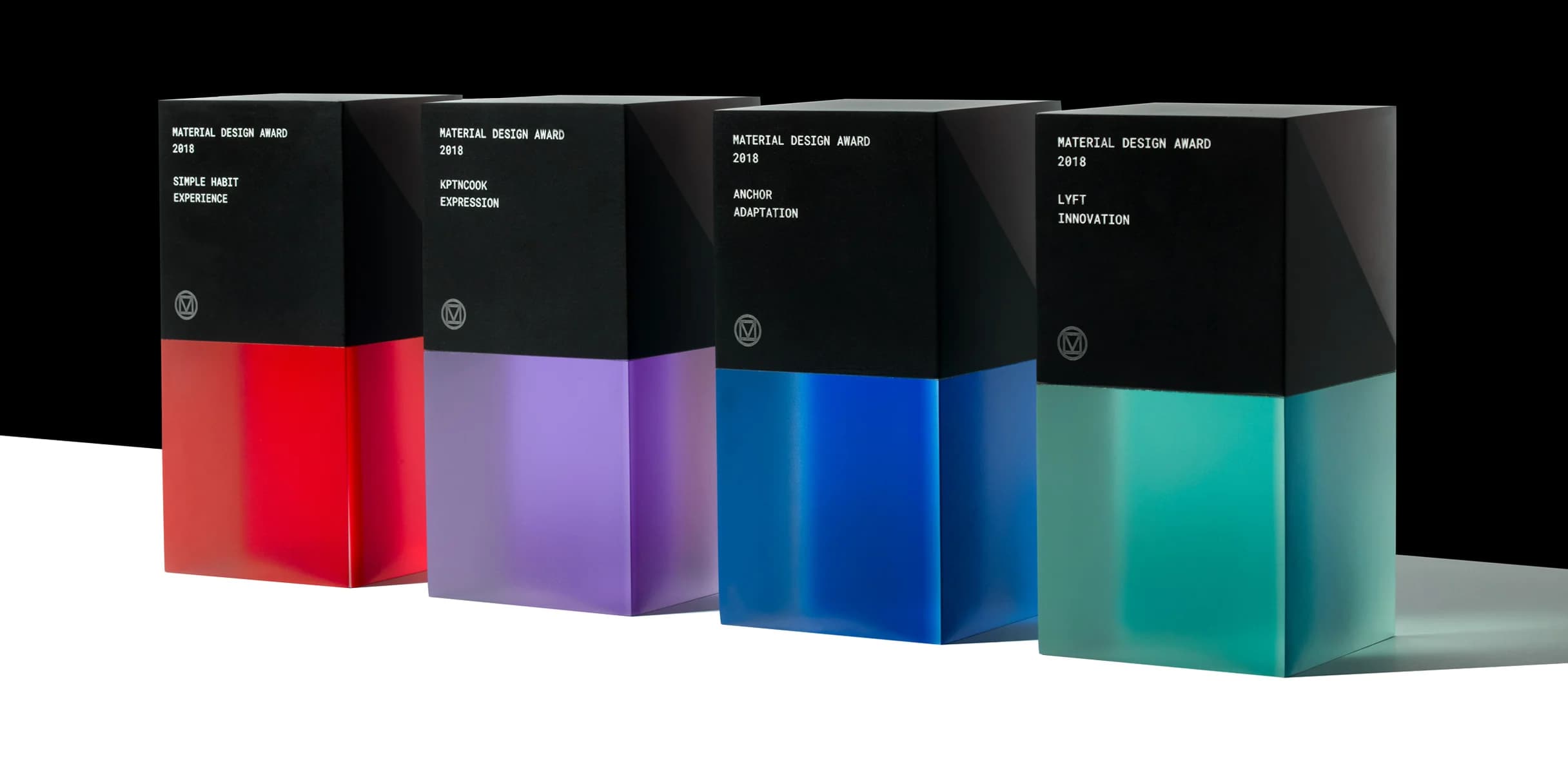 Four Material Design Awards stand on a white surface against a black backdrop. Each acrylic and matte black award features the category and winner's name.