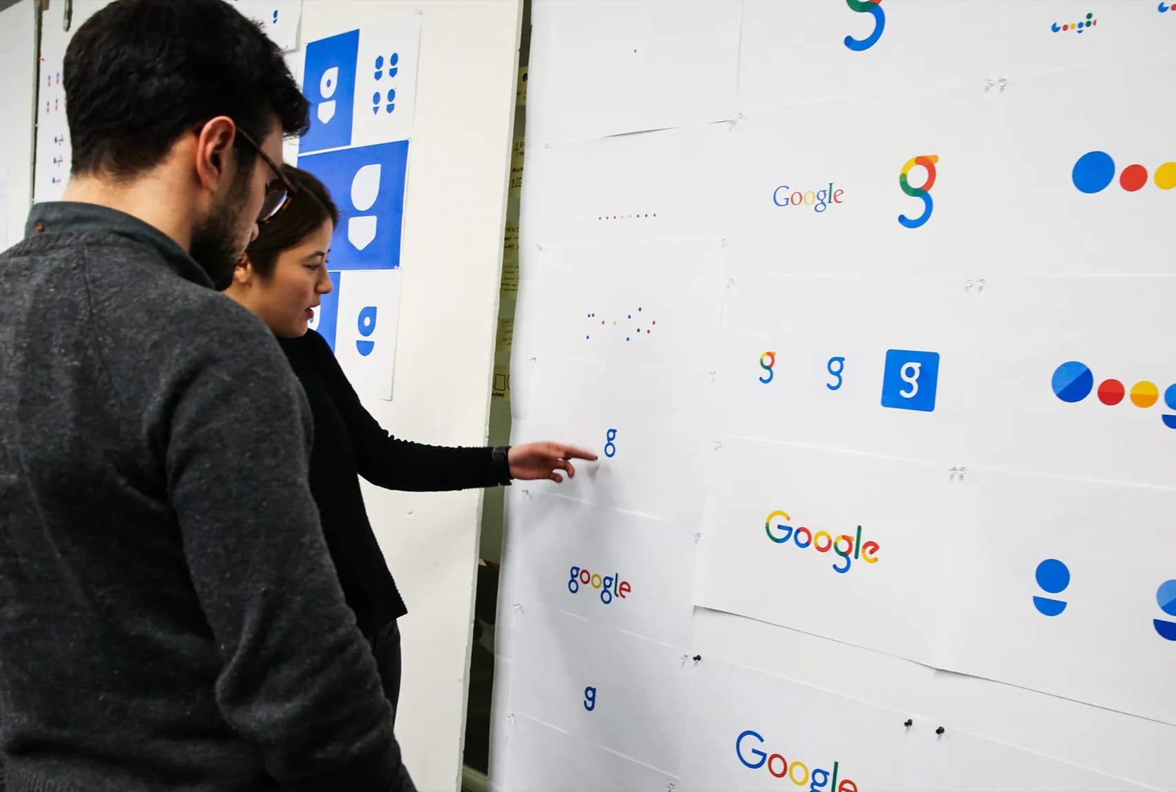 Two people review printed drafts of the Google logo, exploring variations in color, typeface, and composition.