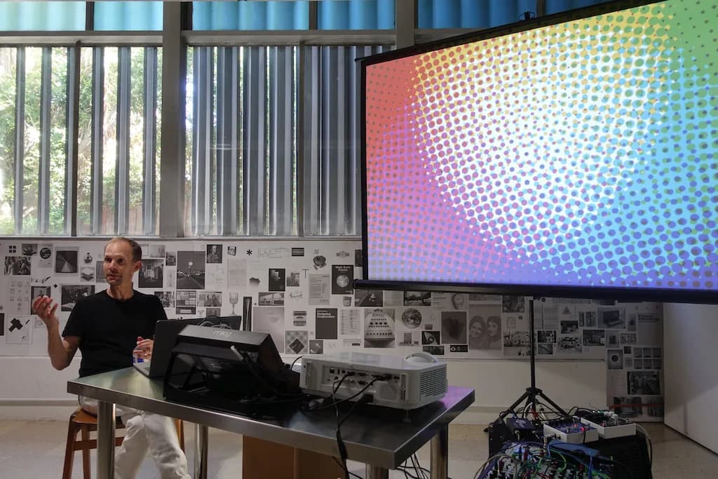 David Reinfurt presents comfortably from a chair next to a large presentation screen.