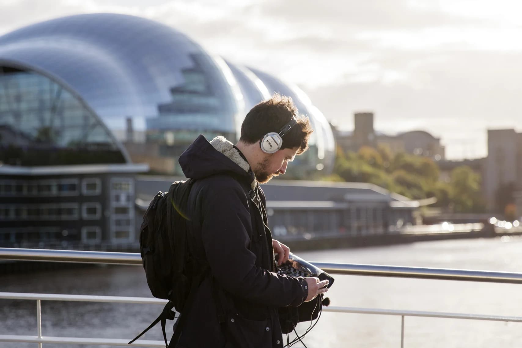 Man with headphones and backpack adjusts a sound mixer while standing on a bridge. A domed building and river are in the background.