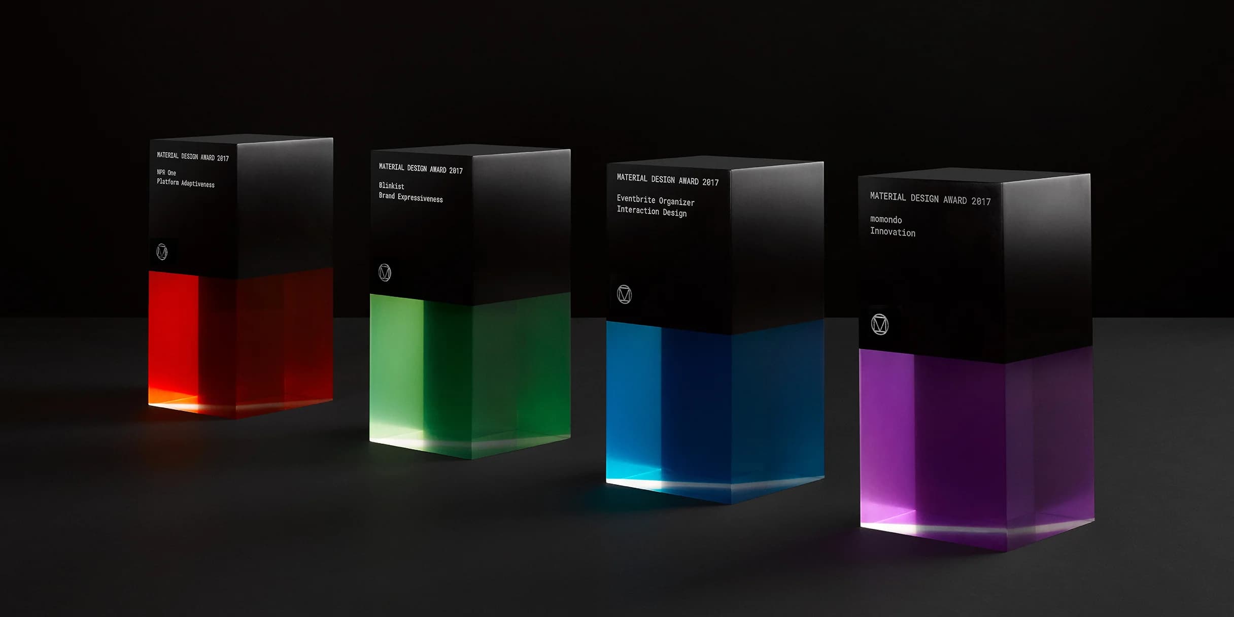 Four Material Design Awards stand in a row. Each black cube features a colorful, translucent base: red, green, blue, and purple.