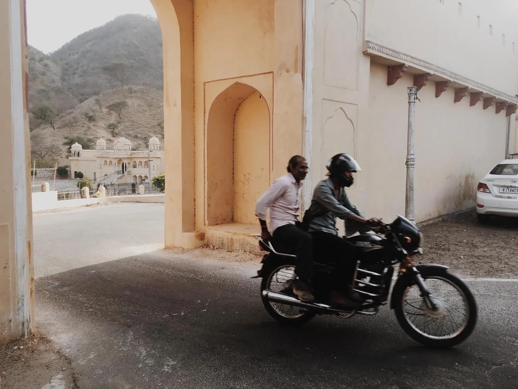 Two men ride a motorcycle through a tall, arched gateway.