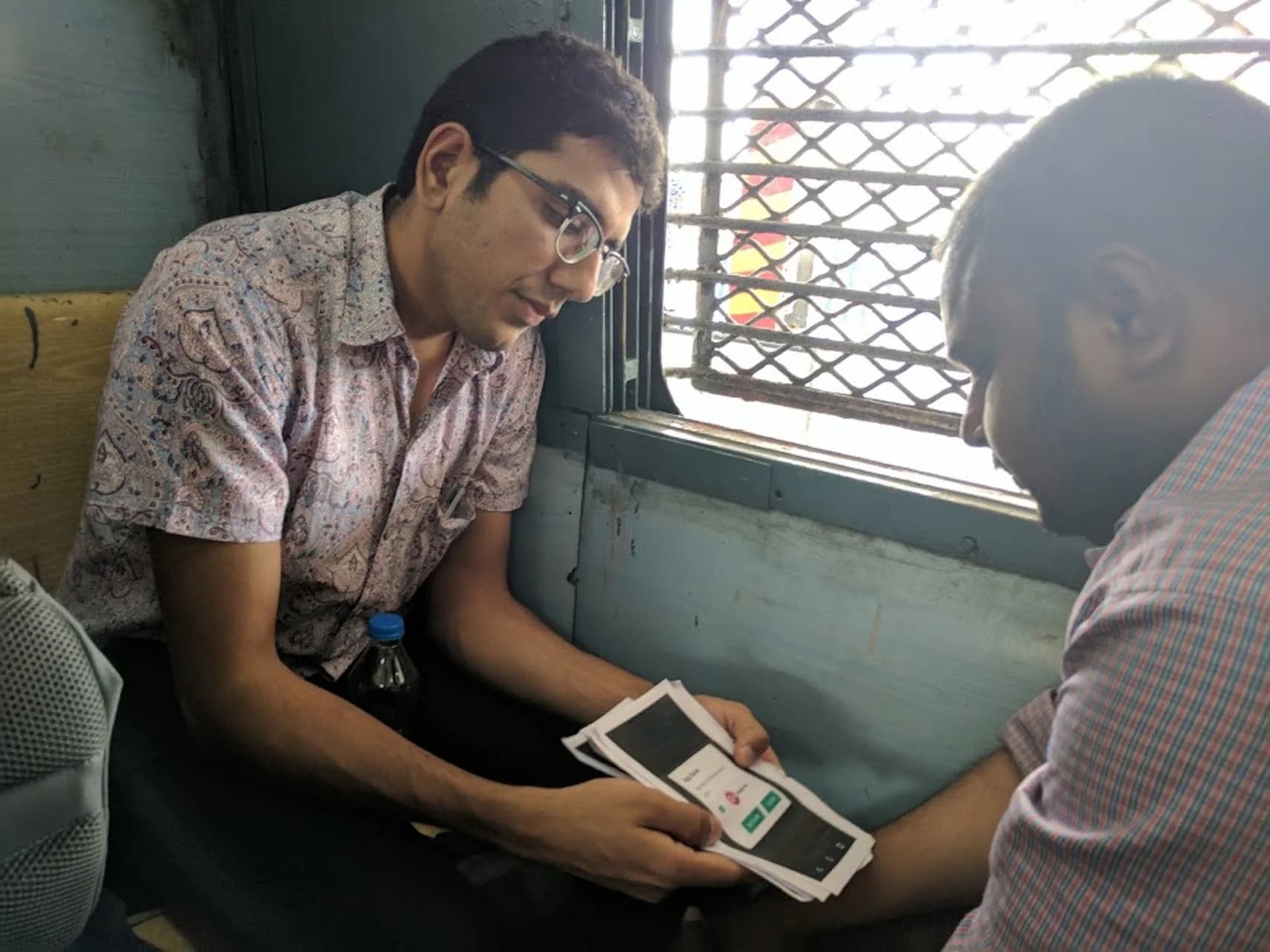 Two men sit on a train. One shows printed app screens to the other, who looks at them.
