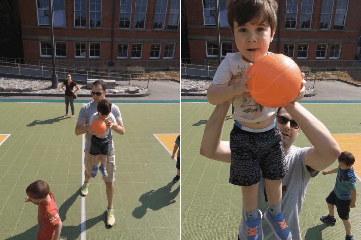 Dad helps their toddler shoot hoops on a sunny basketball court.