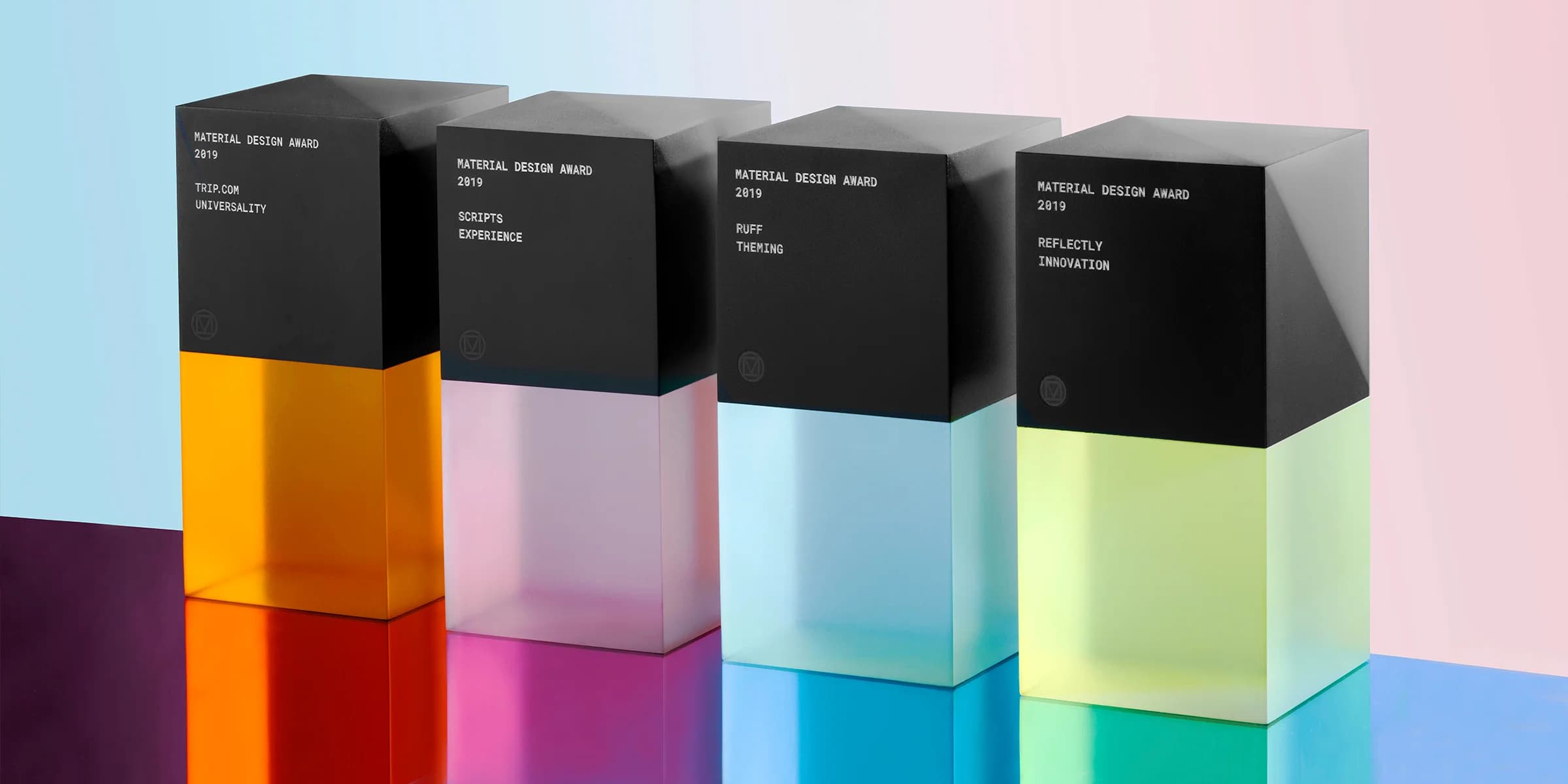 Four Material Design Awards, each a frosted translucent block with a dark top section, stand on a reflective surface. The awards glow orange, pink, light blue, and lime green.