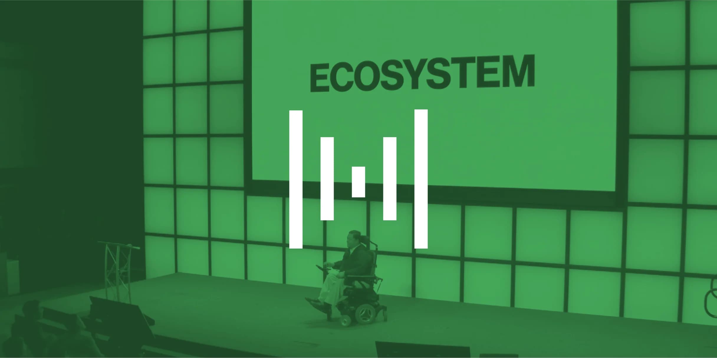 A speaker in a wheelchair presents about "ECOSYSTEM" projected on a large green screen.