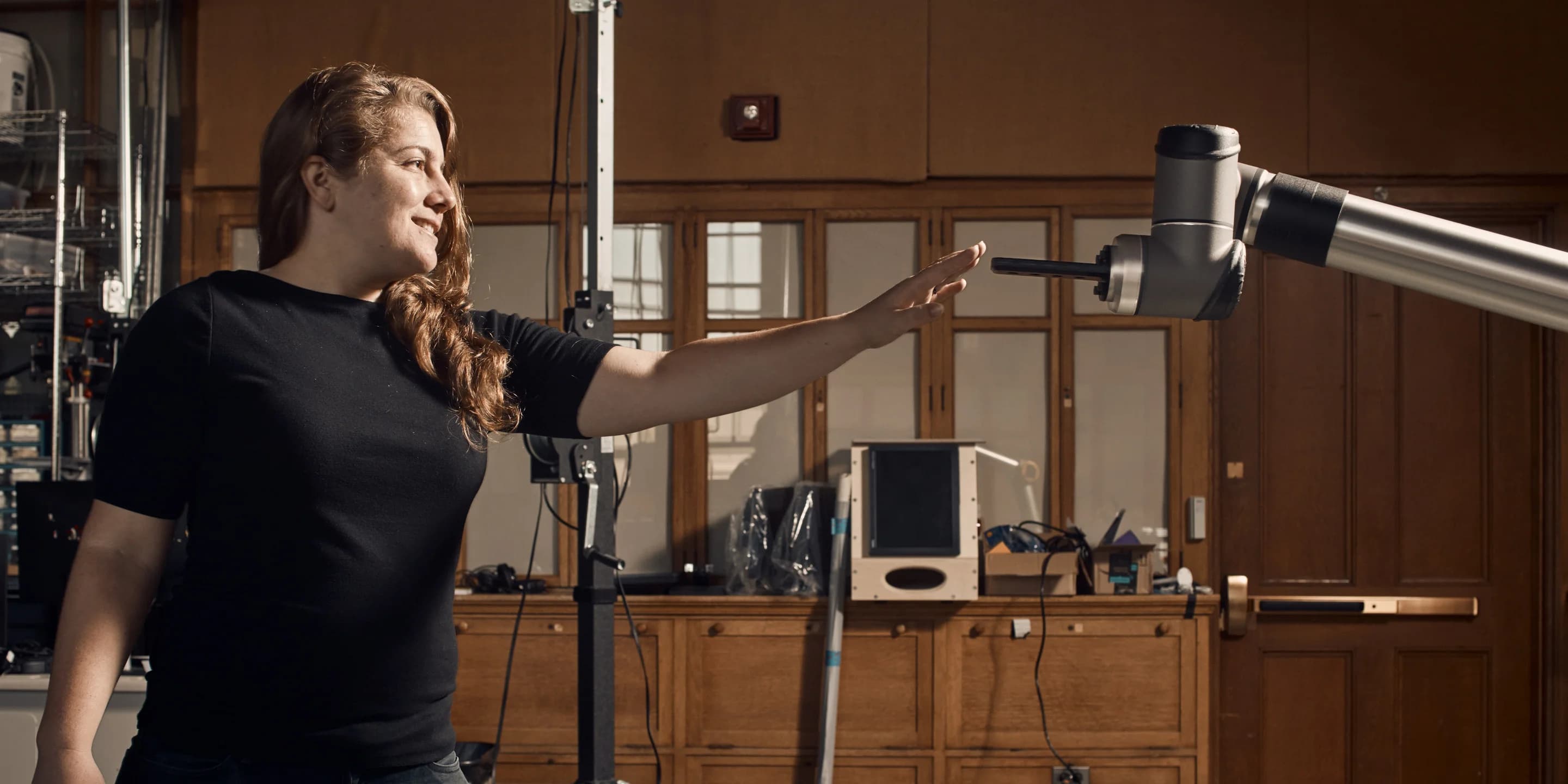 A woman reaches toward a robotic arm extending from the right. She smiles slightly, anticipating its movement.