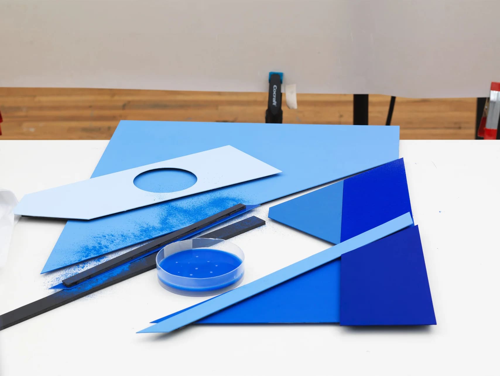 Blue geometric paper cutouts, pigment powder, and a petri dish with blue liquid sit on a white surface.