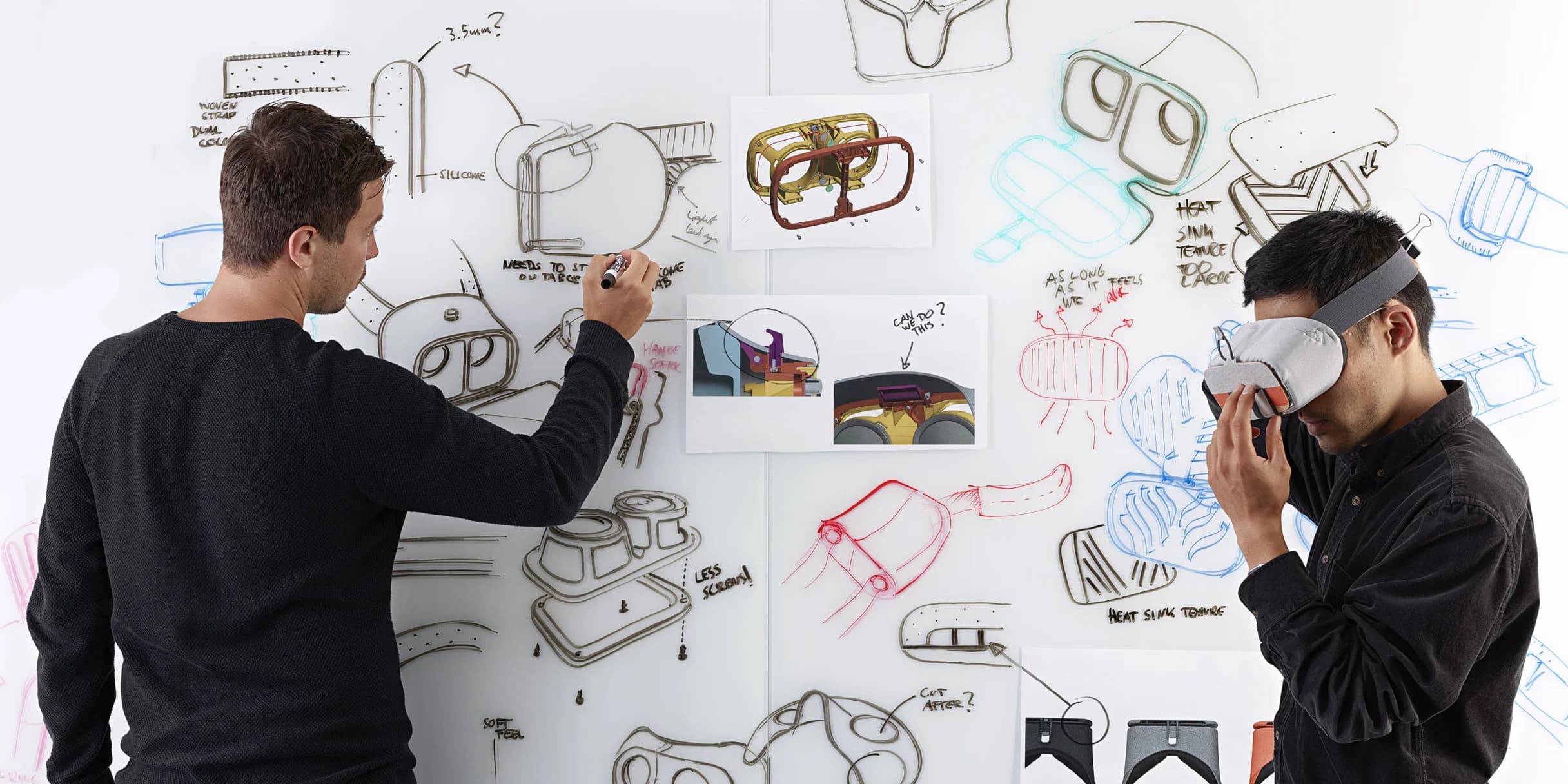 Two men collaborate on VR headset design. One draws diagrams on a whiteboard while the other tests a prototype.