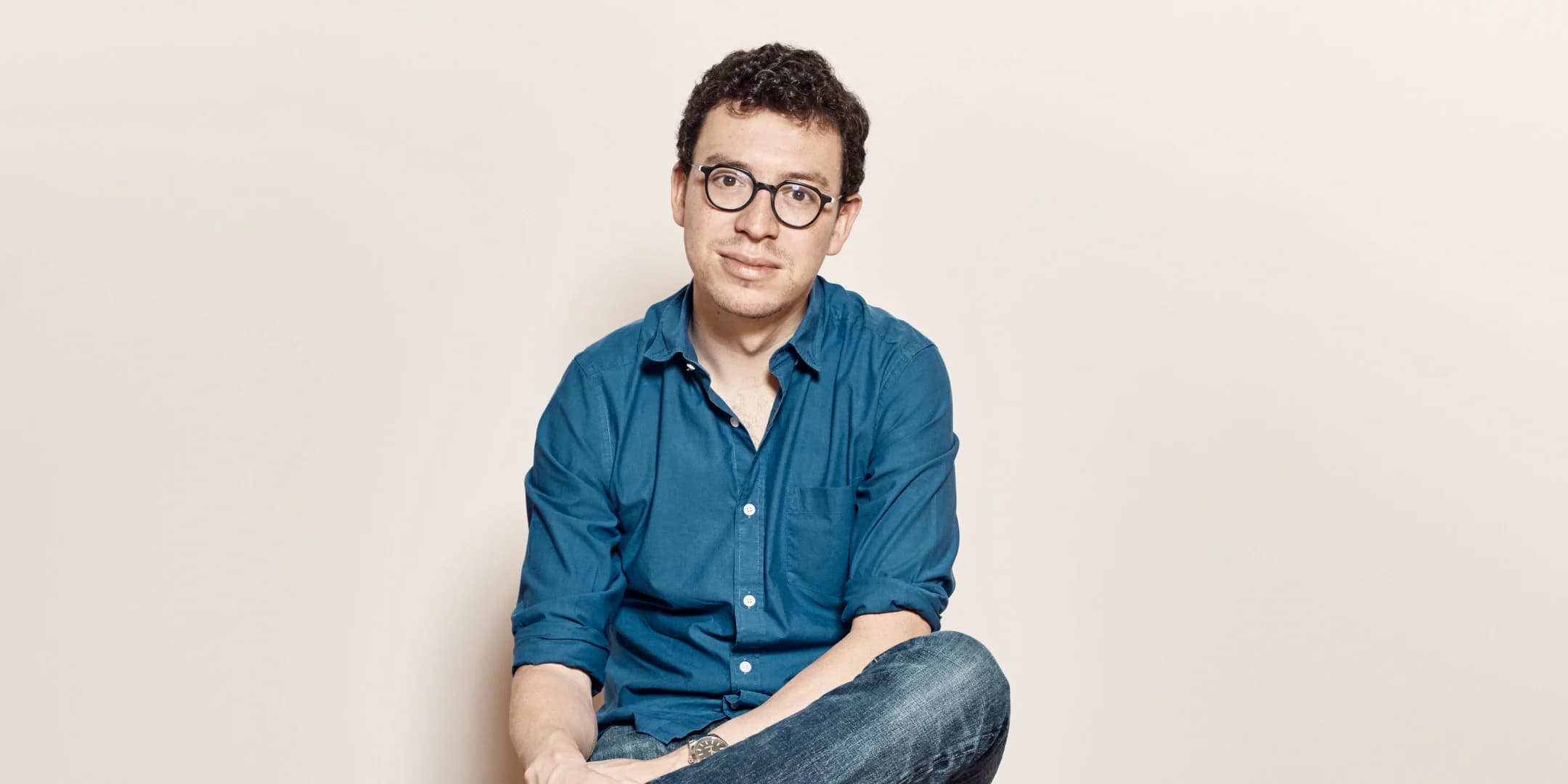 Headshot of a man with glasses, a dark blue button-down shirt, and jeans, sitting with legs crossed, against a cream background.