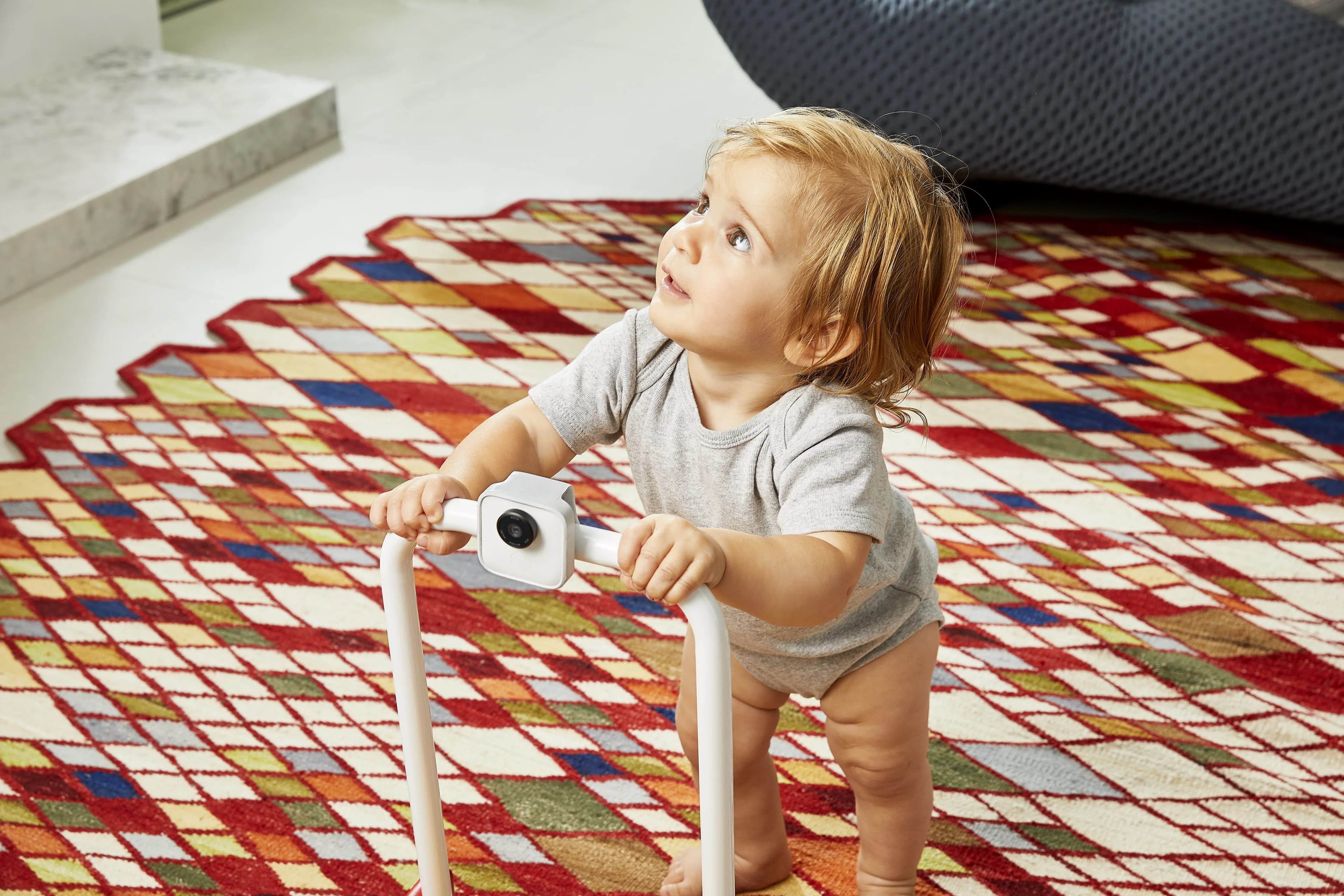 Curious toddler looks up while using a baby walker with a built-in camera. A colorful, patterned rug covers the floor.