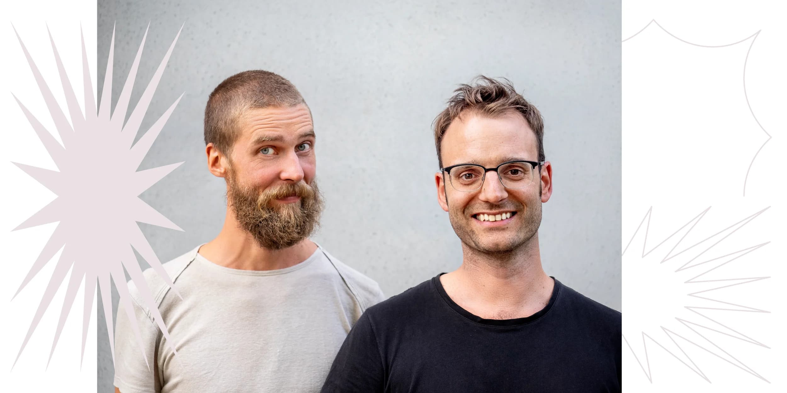 Two men stand shoulder to shoulder against a gray wall. The man on the left has a beard and wears a light gray t-shirt. The man on the right wears glasses and a black t-shirt. Both are smiling.