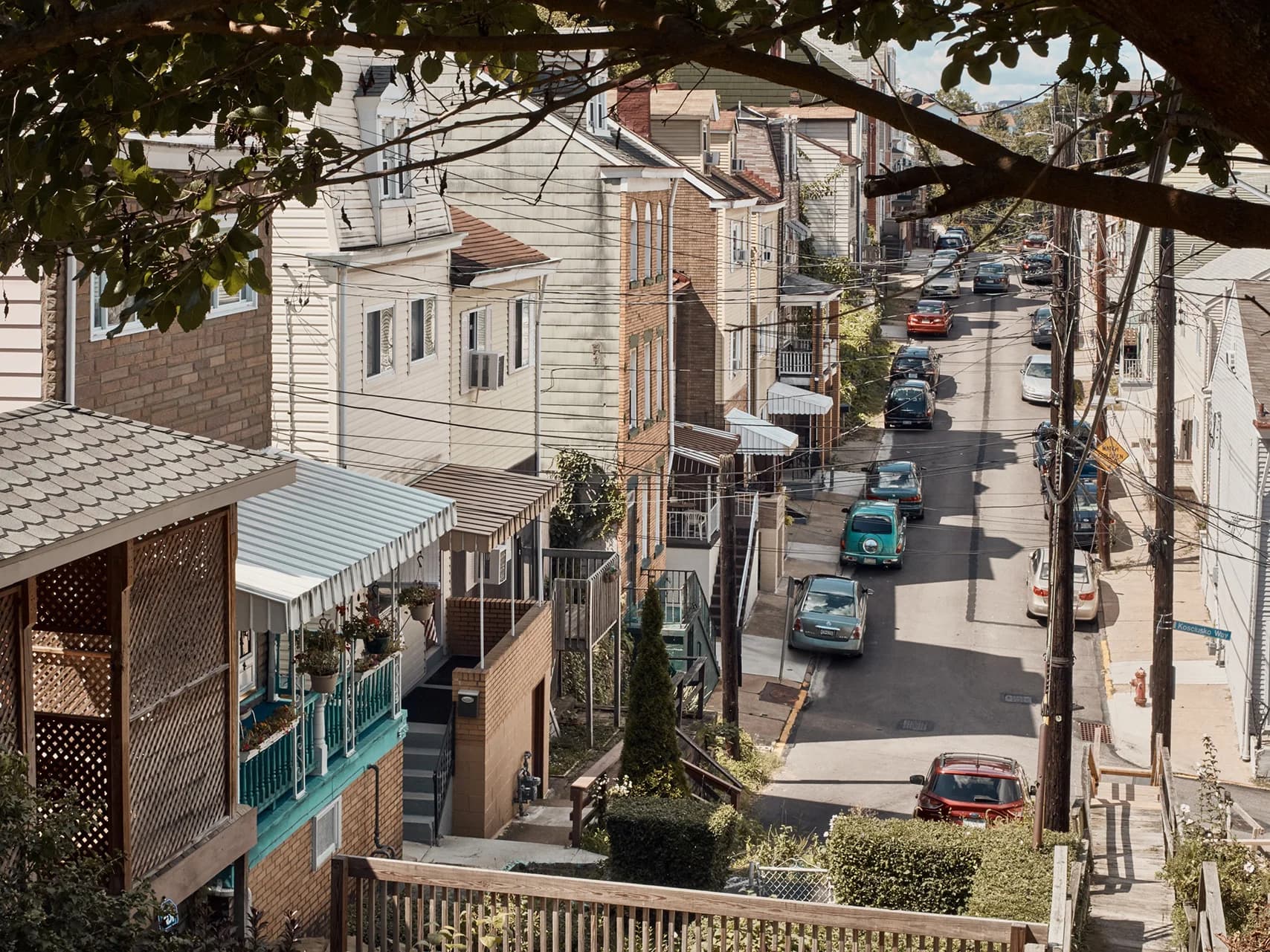 Cars park along a narrow, sloped street lined with colorful houses. Tree branches and powerlines overhang the road. A wooden fence frames the foreground.