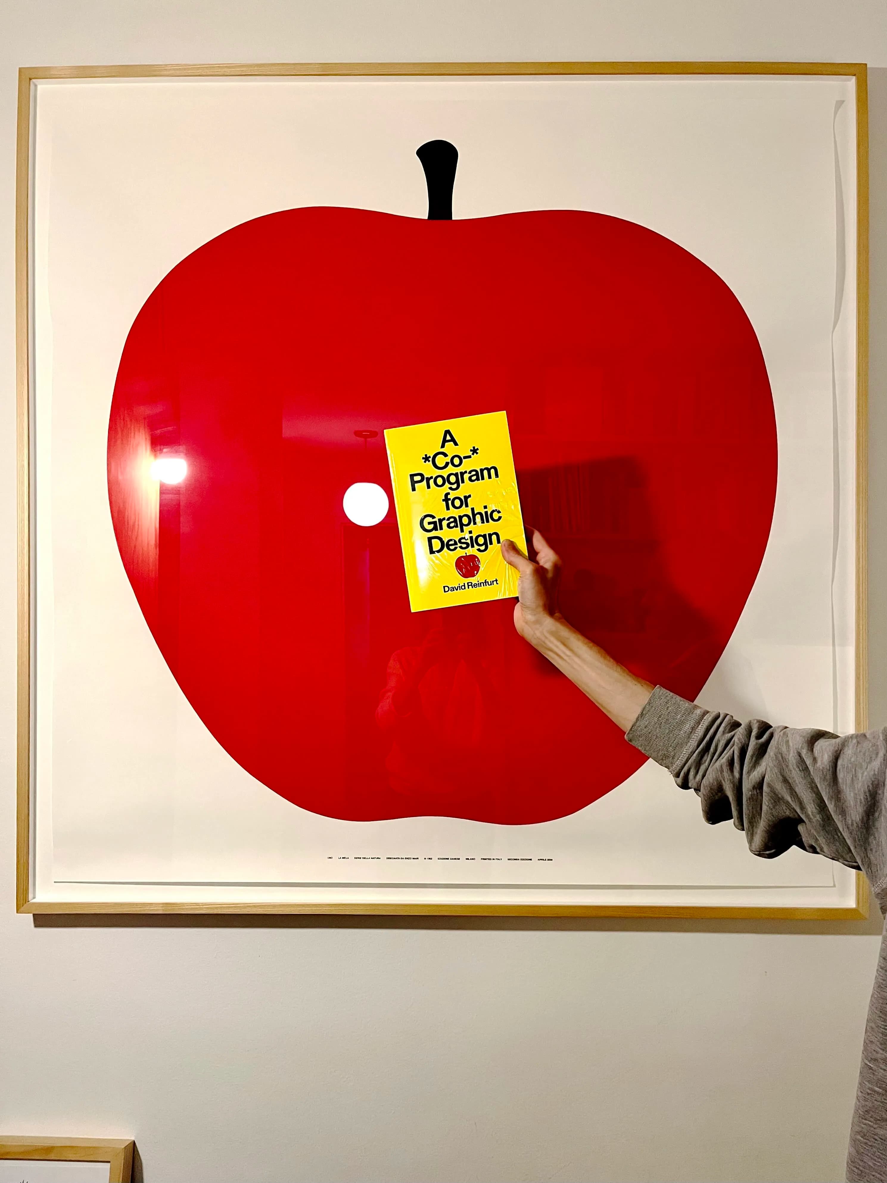 Someone holds up a copy of David Reinfurt’s book in their hand in front of a frame print of La Mela by Enzo Mari.